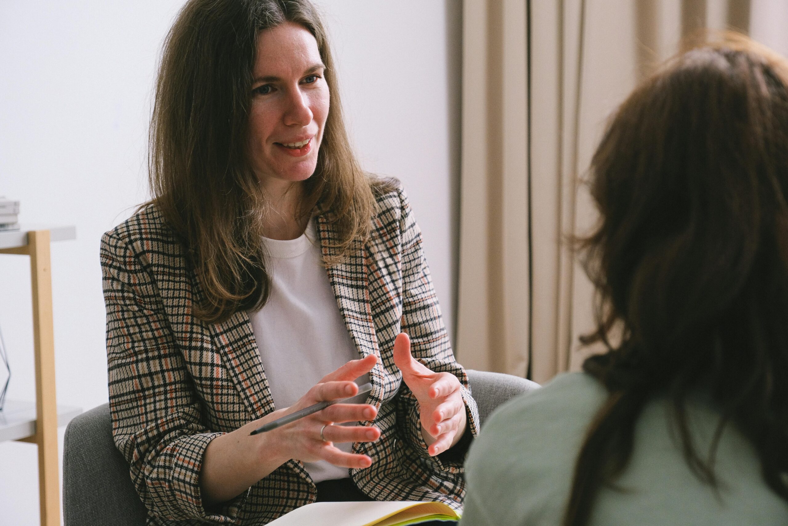 Pensive woman psychologist with brown hair in stylish clothes sitting and talking with unrecognizable female in light room